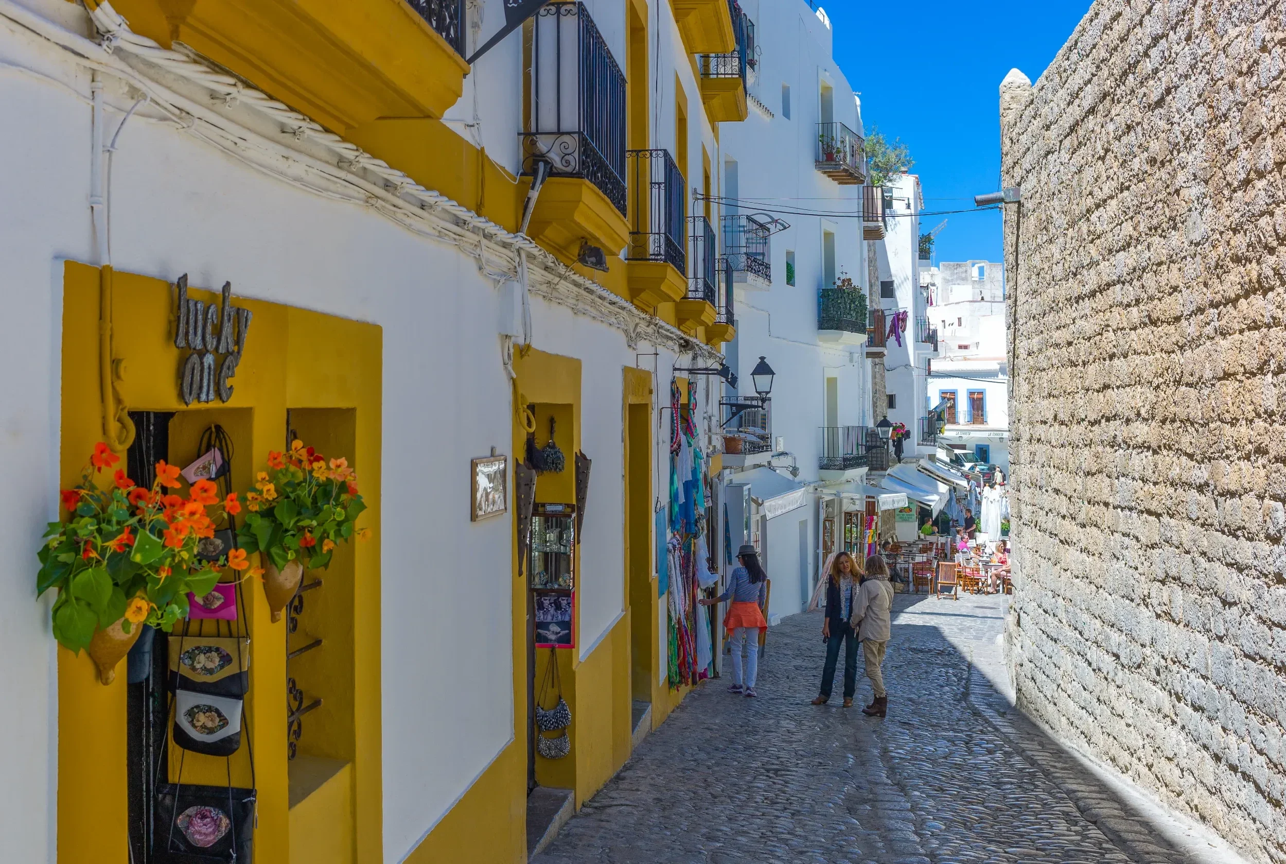 Calle estrecha y empedrada con tiendas y turistas en Ibiza, España. Fachadas blancas con detalles amarillos, balcones con rejas negras y macetas con flores naranjas. Al fondo, más tiendas y personas paseando. Cielo despejado.