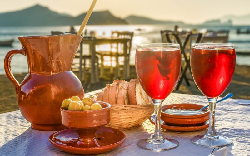 Mesa de restaurante al aire libre junto a la playa, con jarras y copas de sangría, aceitunas y pan en primer plano, capturando el ambiente relajado y pintoresco de una comida en Ibiza al atardecer.
